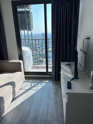 Una habitación moderna con un sofá gris, un escritorio pequeño y vistas de la ciudad a través de un ventanal con cortinas negras.