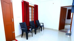 A room with a white tiled floor, a wooden door, and a window with red curtains. There are four black chairs arranged in the room. - Tea Haze Cottage Munnar (Anachal)