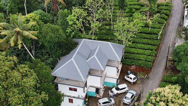 A house with a sloped roof, surrounded by lush greenery and a road. - Tea Haze Cottage Munnar (Anachal)