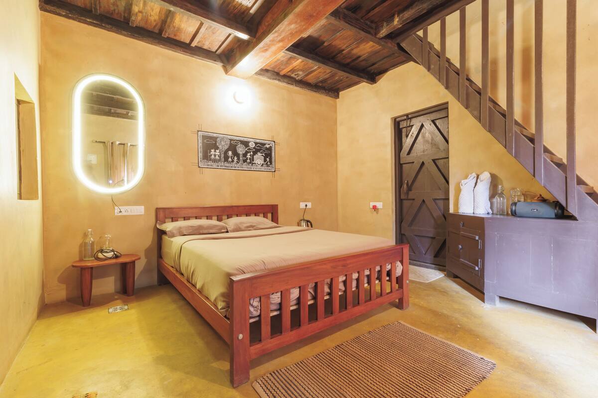A bedroom with a wooden bed, a bedside table with a lamp, a wall-mounted mirror, and a staircase leading to another room.