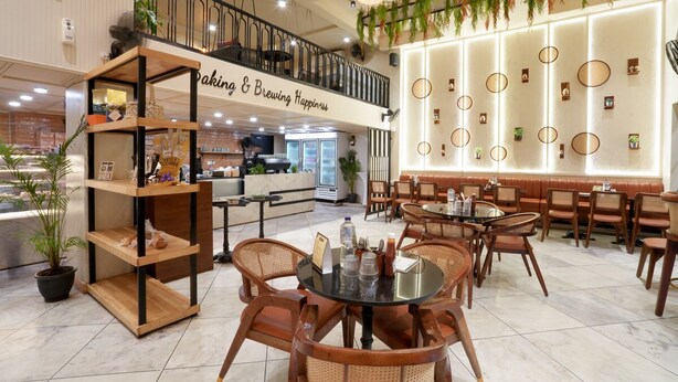 A modern cafe with wooden furniture, a sign reading "Baking & Brewing Happiness," and a balcony with plants. - Hotel Square 36 (Kota)