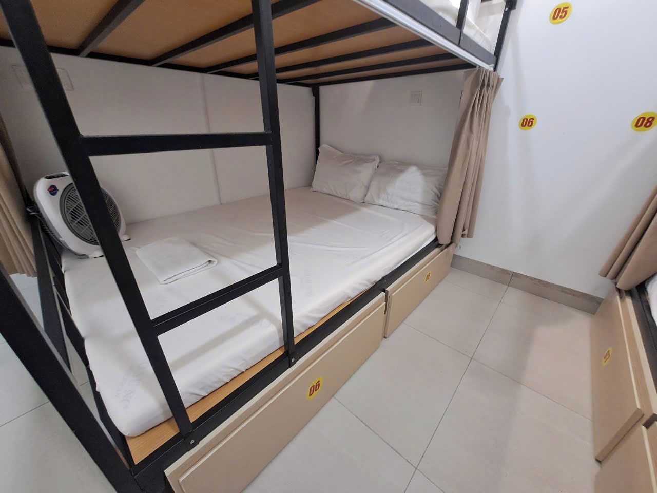 Una cama individual en literas con un ventilador, un mueble pequeño y una cortina.
