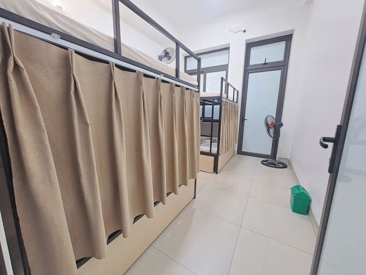 A hotel room with a bunk bed, a window with blinds, a door, and a green trash bin.