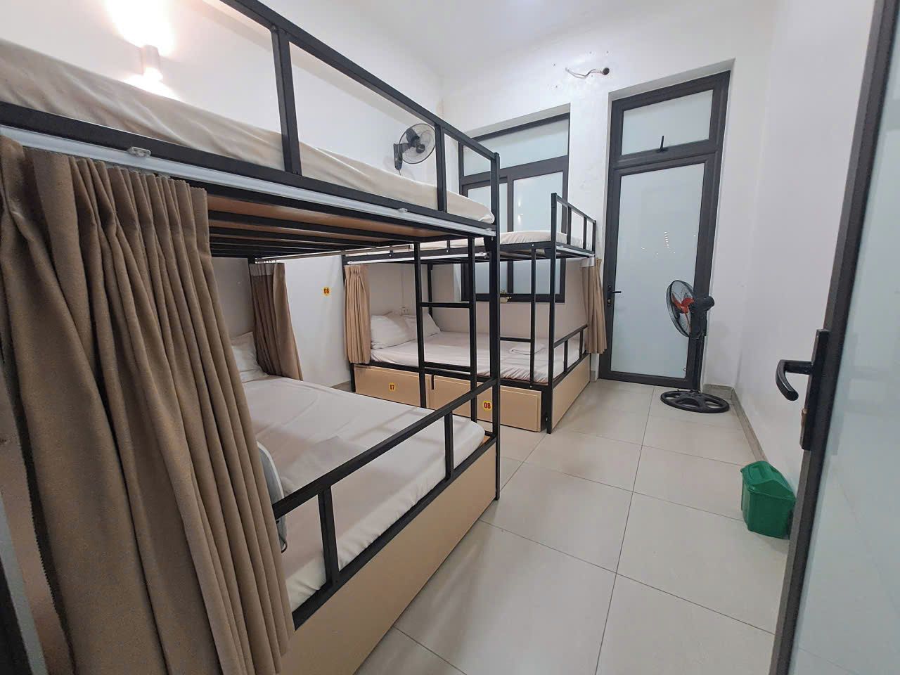 A dormitory room with bunk beds, a fan, a door, and a green trash bin.