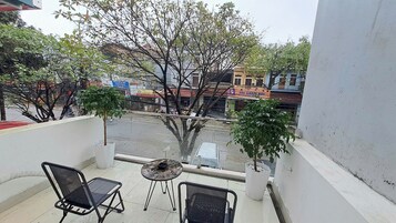 Varanda com duas cadeiras e uma mesa pequena, com vista para uma rua com edifícios e árvores.