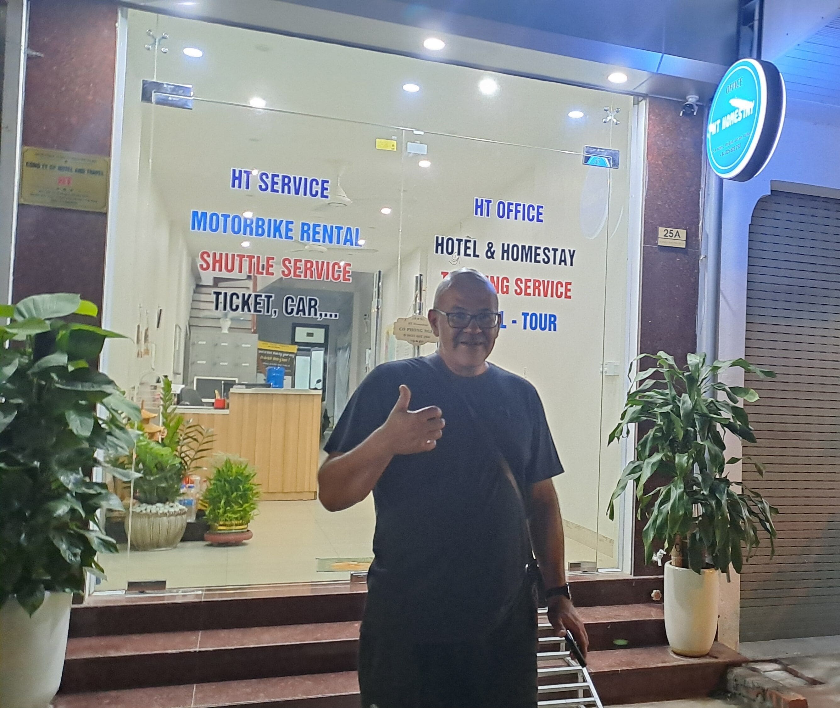 A man standing in front of a building with signs for motorbike rental, shuttle service, hotel & homestay, and tour services.