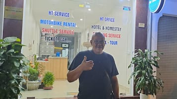 A man standing in front of a building with signs for motorbike rental, shuttle service, hotel & homestay, and tour services.