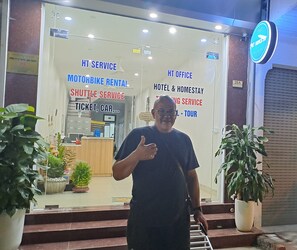 A man standing in front of a building with signs for motorbike rental, shuttle service, hotel & homestay, and tour services. - HT Airport Inn (Hanoi)