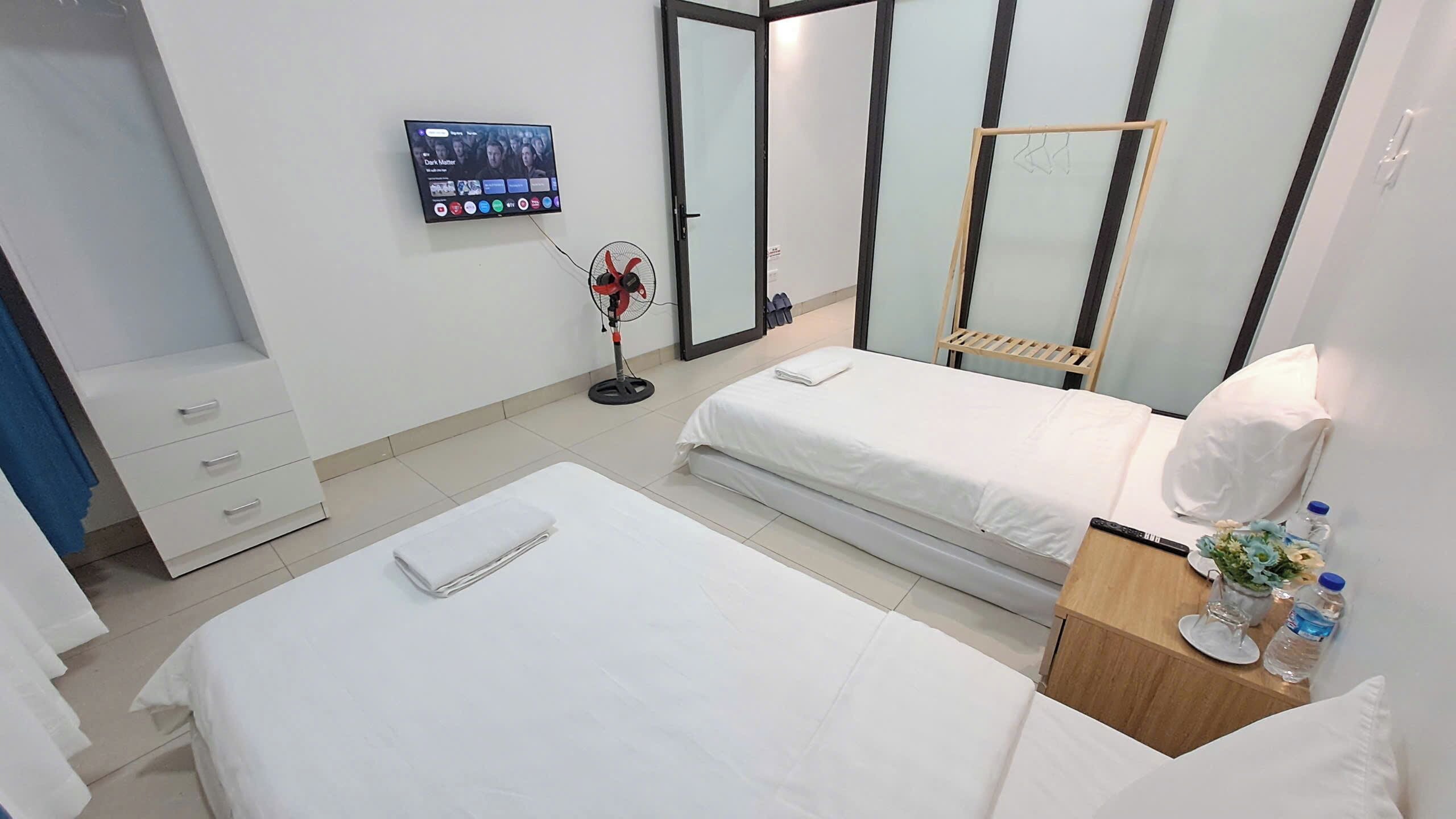 A hotel room with two beds, a TV mounted on the wall, a fan, and a wooden nightstand with a water bottle and glasses.
