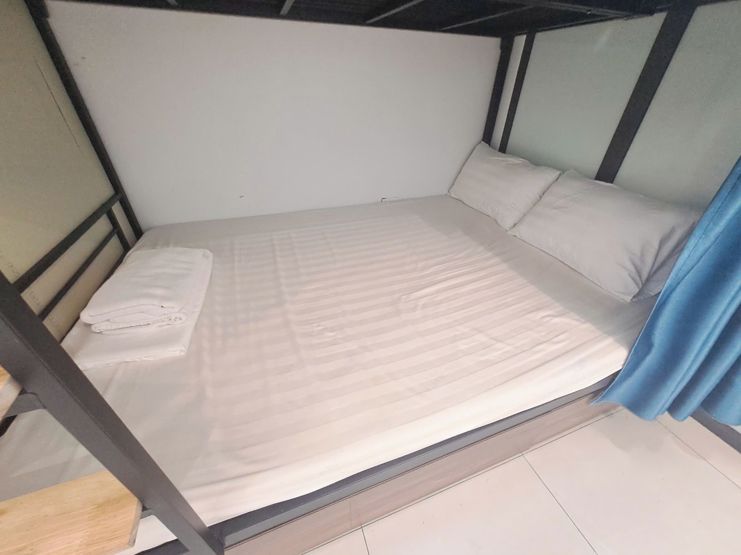 Una cama individual con colchón y almohadas blancas, una manta doblada y una cortina azul.