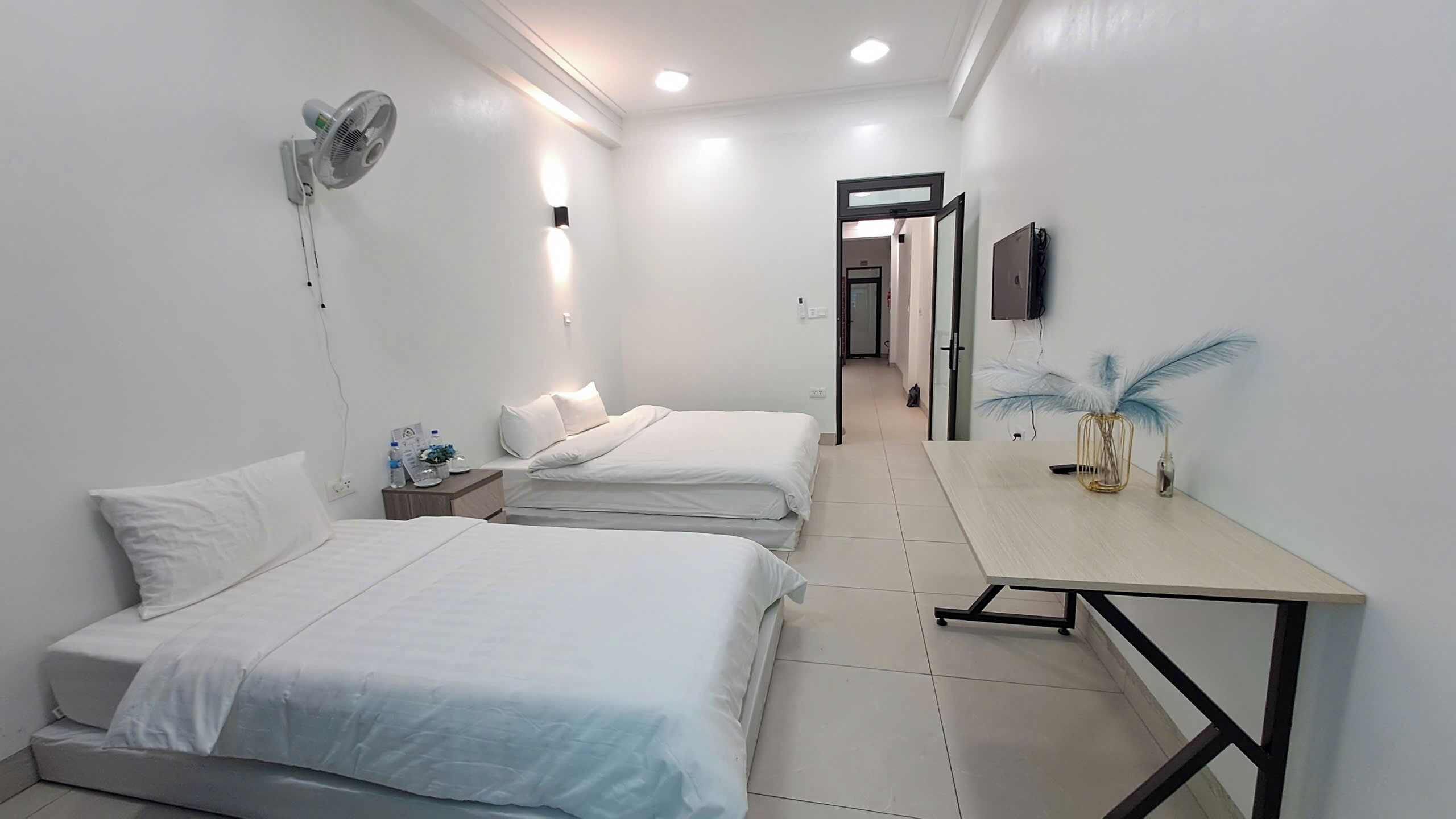 A hotel room with two beds, a desk, a fan, and a TV on the wall.