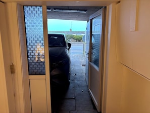 A narrow hallway with a car parked on the left, a door with a frosted glass panel on the right, and a clear sky visible through the window.