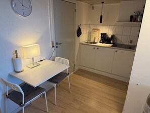 Une cuisine compacte avec un plan de travail blanc, un évier et une machine à café. On y trouve également un petit coin repas avec une table et des chaises.