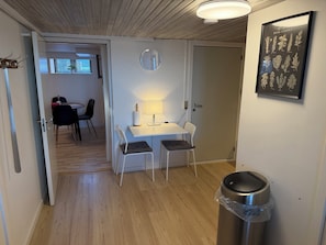 A room with a wooden ceiling, a white desk with a lamp, two chairs, a wall clock, a framed botanical illustration, and a trash bin.