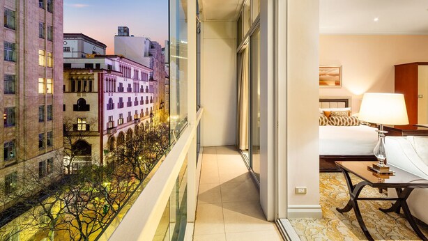 Habitación de hotel con un ventanal grande que da a un paisaje urbano, una cama con cabecera blanca y una mesita con lámpara.