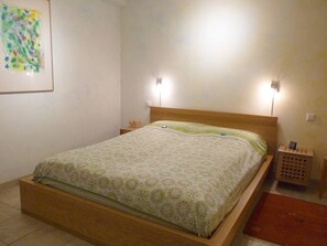 Un lit simple avec un cadre en bois, une couvre-lit à motifs, deux lampes de chevet, une petite table de chevet avec une lampe et un tableau encadré au mur.
