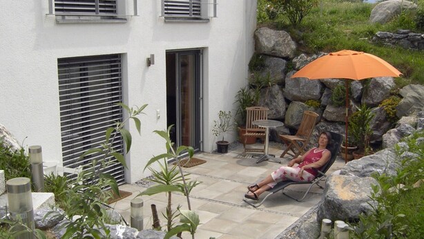 A woman relaxing on a chair under an umbrella in a garden area with potted plants and a stone wall.