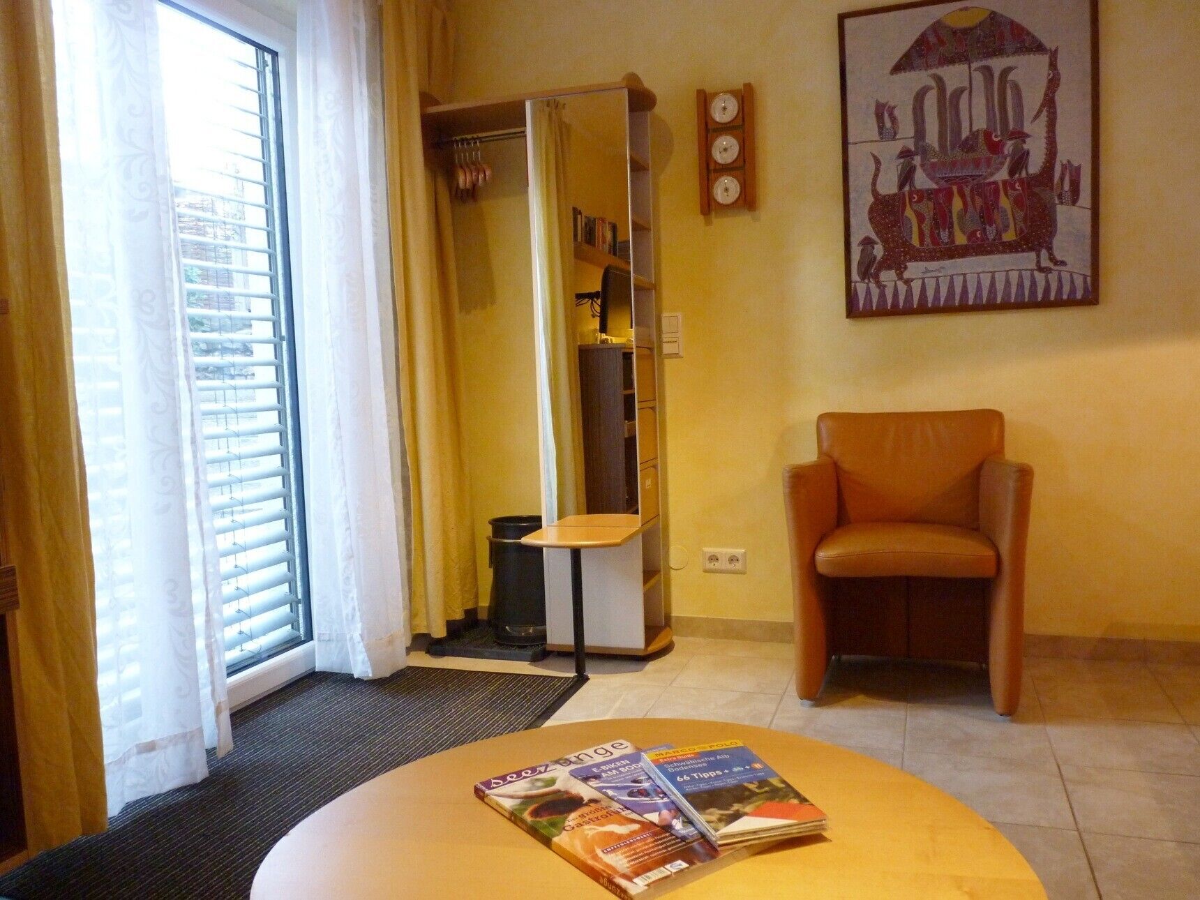 Une chambre d’hôtel avec une porte-fenêtre coulissante, une petite table avec des magazines, une chaise, un porte-manteau et une horloge murale.