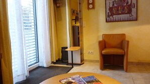 A hotel room with a sliding glass door, a small table with magazines, a chair, a coat rack, and a wall clock.