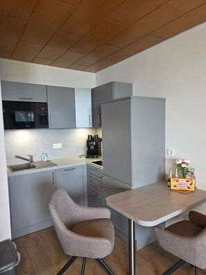 A compact kitchen with grey cabinetry, a built-in microwave, and a coffee machine. There are two chairs and a small table with a box of Knusper Schoklis cookies.