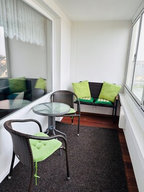Una habitación de esquina con una mesa de vidrio, sillas de mimbre, un banco acolchado y vistas a través de una ventana con cortinas de gasa.