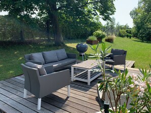 A deck with a grey sofa, a small table, and a chair, surrounded by greenery and trees. - Holiday Apartment 'Bodenseeloft H E 66' (Kressbronn am Bodensee)