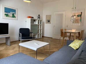 Una sala de estar con sofá, televisor, estantería, mesa de comedor y sillas, además de una puerta.