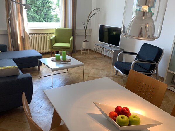 A modern living room with a sofa, a dining table, a TV, and a lamp. - Apartment 'Bodenseeloft HE 66' (Kressbronn am Bodensee)