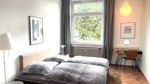A bedroom with a bed, a desk, a chair, a radiator, and a window with curtains. - Apartment 'Bodenseeloft HE 66' (Kressbronn am Bodensee)