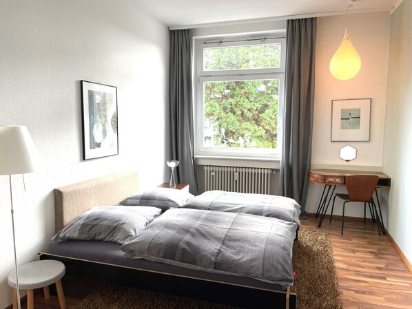 A bedroom with a bed, a desk, a chair, a radiator, and a window with curtains. - Apartment 'Bodenseeloft HE 66' (Kressbronn am Bodensee)