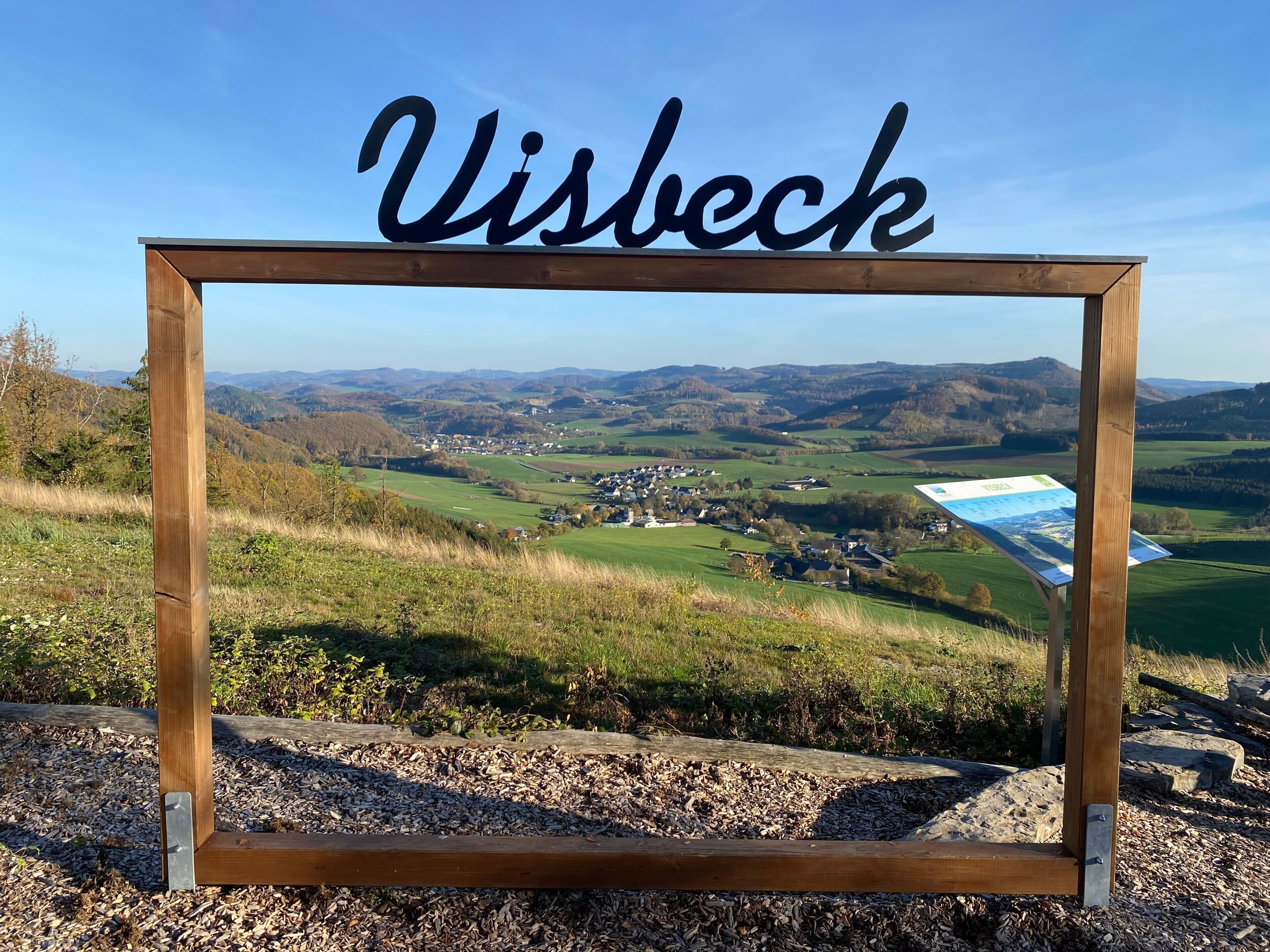 Un letrero de madera con la palabra 'Visbeck', con vistas a un paisaje pintoresco de colinas y un pueblo pequeño.