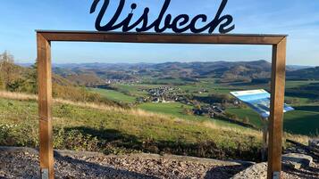 Un letrero de madera con la palabra 'Visbeck', con vistas a un paisaje pintoresco de colinas y un pueblo pequeño.