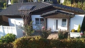 Una casa con techo inclinado, paneles solares, una antena parabólica y un porche pequeño con un banco.