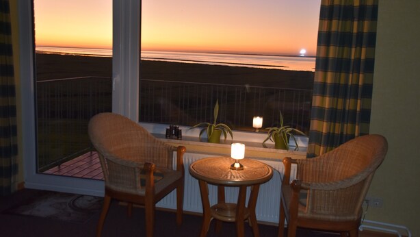 Ein Zimmer mit zwei Korbstühlen, einem kleinen runden Tisch mit Kerze und Blick auf den Sonnenuntergang über dem Meer durch große Fenster.