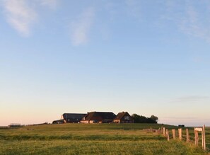 A rural landscape with a large barn and a smaller house, surrounded by open fields and a wooden fence.