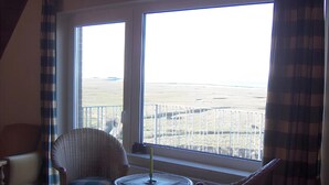 Ein Zimmer mit großem Fenster, Korbsessel, rundem Tisch und Heizkörper.