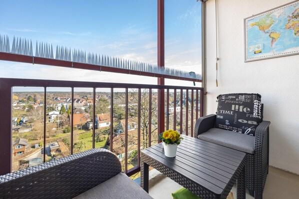 Un balcon donnant sur un quartier résidentiel, une chaise en osier avec un coussin, une petite table avec une fleur et une carte du monde accrochée au mur.
