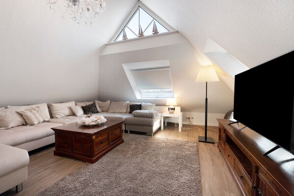 A loft-style living room with a high ceiling, a large window, a flat-screen TV, a sofa, a coffee table, and a lamp.