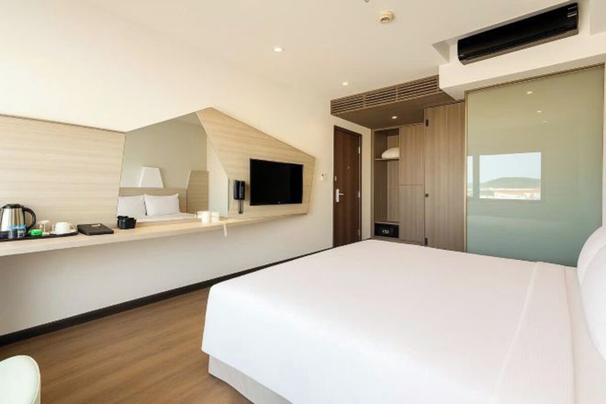 A modern hotel room with a large bed, a flat-screen TV mounted on the wall, a small table with a lamp, and a view of the city through a window.