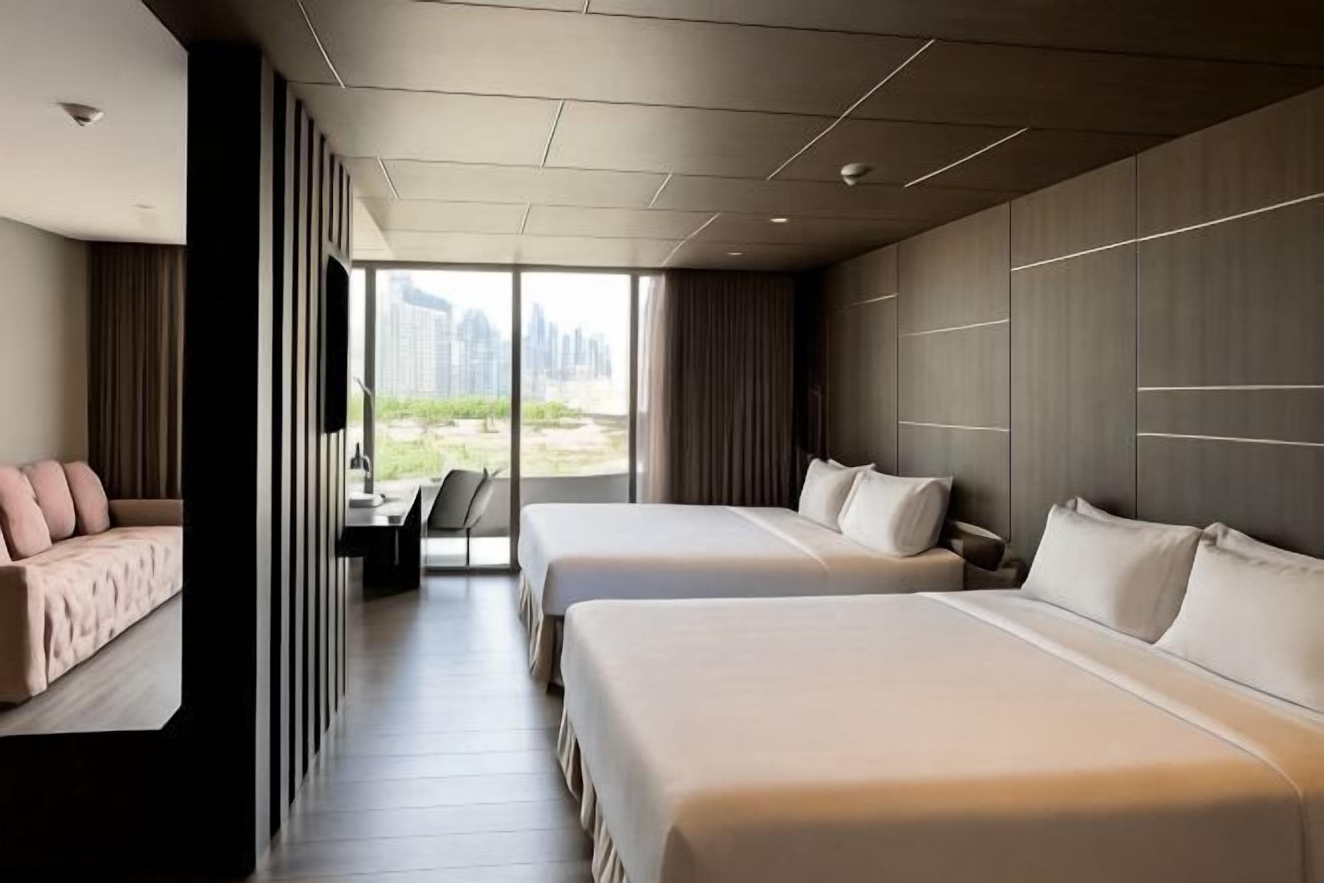 Ein Hotelzimmer mit zwei Betten, einem Sofa, einem Schreibtisch und einem großen Fenster mit Blick auf die Stadt.