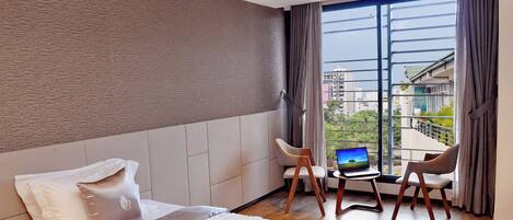 A hotel room with a bed, a desk with a laptop, two chairs, and a large window with a city view.