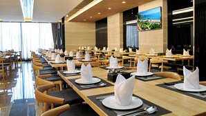 Un comedor amplio con mesas largas dispuestas para varios invitados, con elegantes vajillas y una pantalla grande que muestra una vista panorámica.
