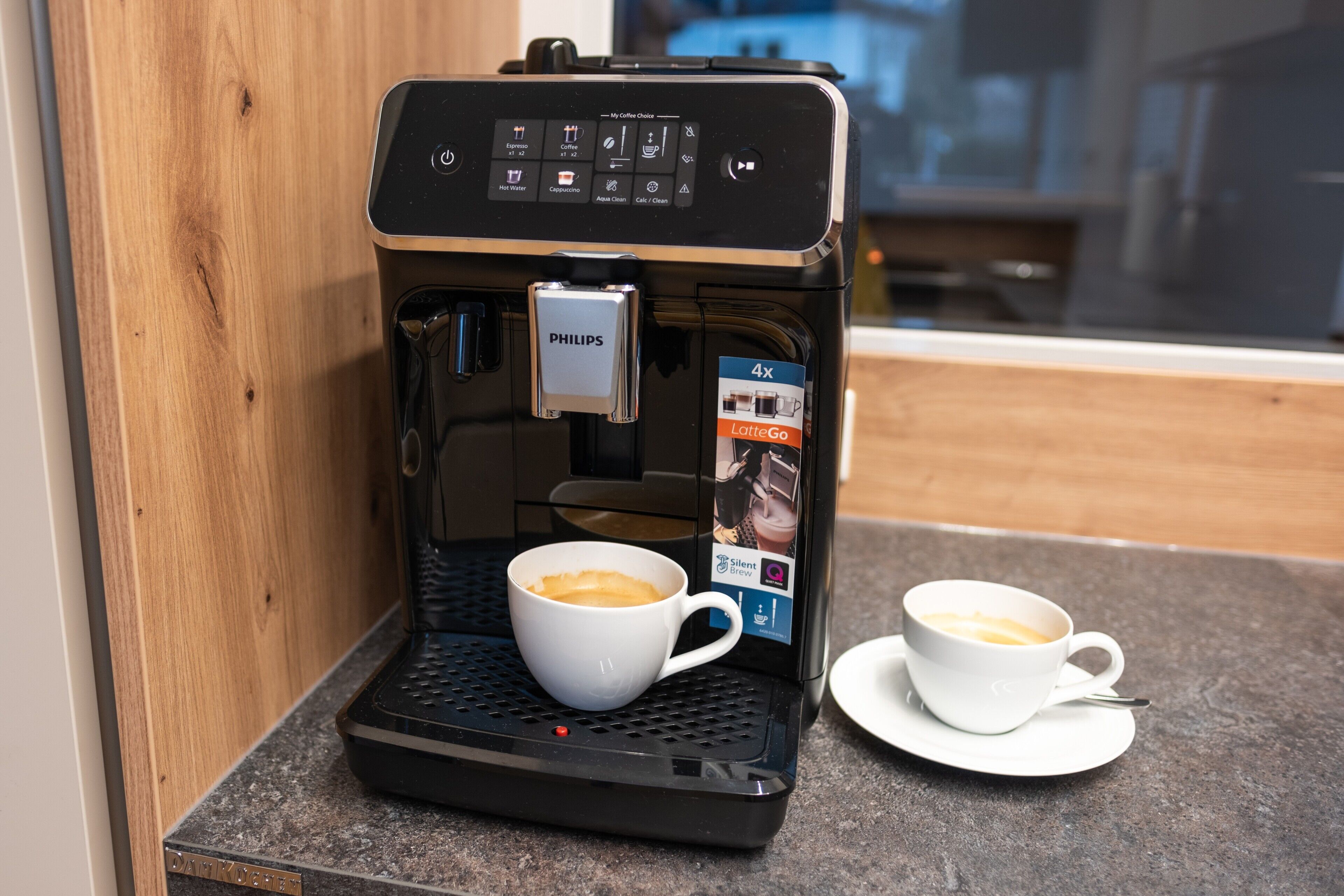 Een koffieapparaat met een kop koffie op een dienblad, een tweede kop op een schoteltje, en een houder voor koffiecups.