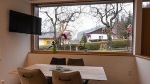 Un comedor con mesa y sillas, un televisor de pantalla plana montado en la pared y una ventana con vistas a árboles y casas.