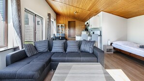 A modern room with a wooden ceiling, a grey sectional sofa with patterned cushions, a wooden bedside table, and a bed with a white mattress.