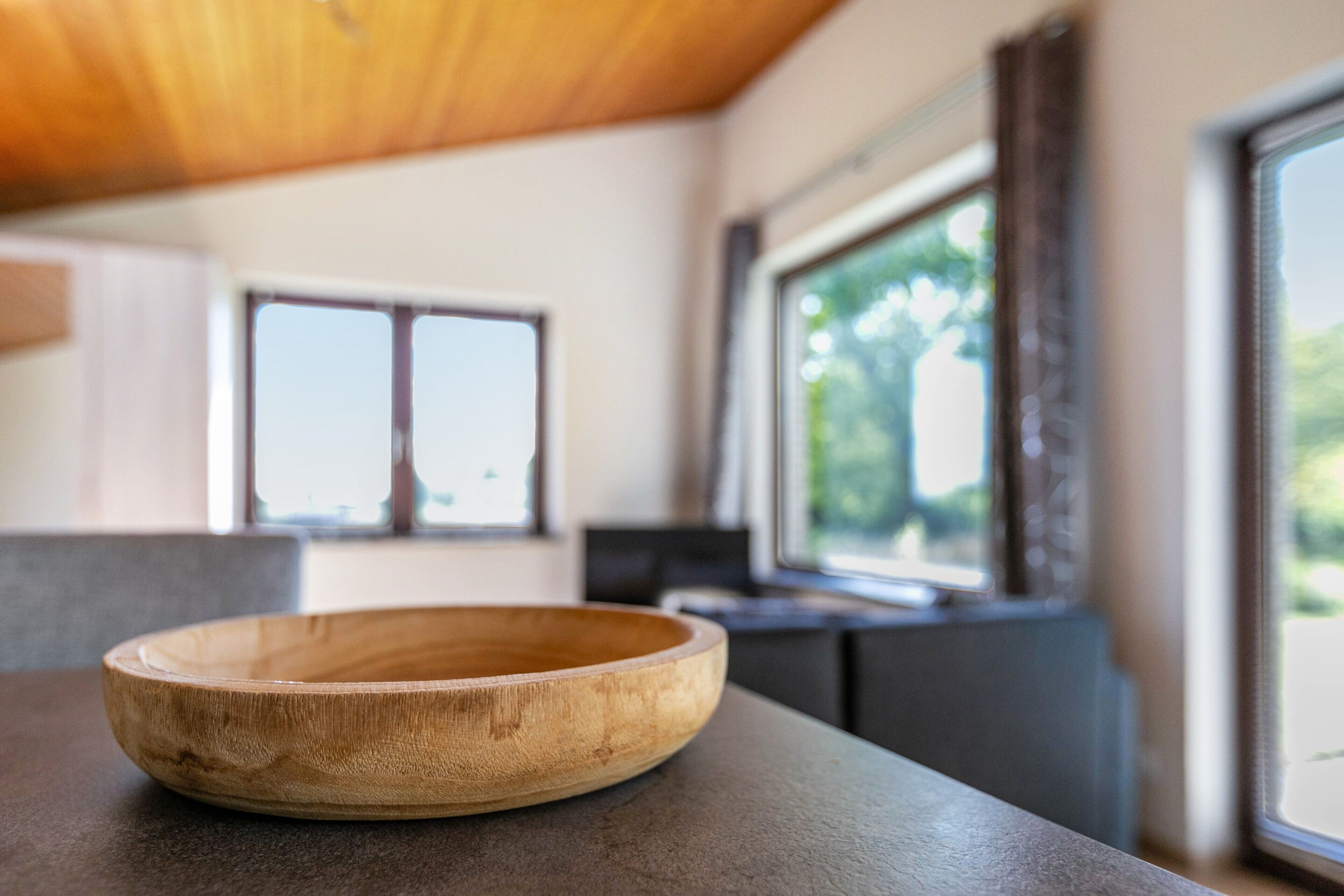 A wooden bowl on a countertop in a modern kitchen with large windows and a view of greenery outside.