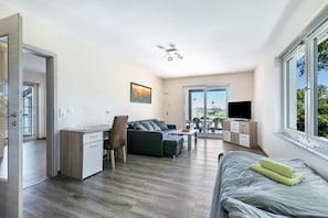 A modern apartment with a kitchenette, a sofa, a bed, a TV, and a large window. - Holiday Apartment 'Wiesenblick 1' with Private Terrace, Private Garden and Wi-Fi (Weddingstedt)