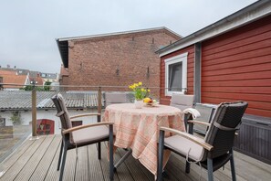 Une terrasse en bois avec une table et des chaises, un vase de fleurs et, au fond, un bâtiment rouge.