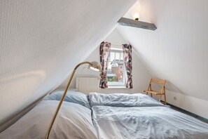 Une chambre avec un plafond en pente, un lit recouvert de linge de lit blanc, une lampe sur pied et une vue sur un immeuble à travers une fenêtre ornée de rideaux à motifs.