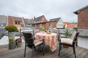 A rooftop terrace with a round table, chairs, potted plants, and a view of surrounding buildings.